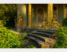 Goldene Statuen und reich verzierte Säulen zieren die Außenfassade eines grünen Pavillons, der von Bäumen und dichter Vegetation umgeben ist. Im Vordergrund führt eine geschwungene Steintreppe durch den sorgfältig gestalteten Garten zur Struktur.