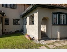 Ein Wohnhaus mit hellem Putz, grünen Fenstereinfassungen und Sprossenfenstern. Im Vordergrund befindet sich ein kleiner Garten mit Rasen, Steinweg und Blumentöpfen, während ein überdachter Eingangsbereich mit Rundbogen und Treppe gut sichtbar ist.