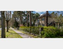 Wohnstraße mit einem Gehweg, Bäumen und einem eingezäunten Garten; dahinter ein zweistöckiges Wohnhaus mit Balkon, umgeben von hohen Kiefern und weiteren Pflanzen.
