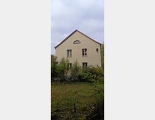 Ein zweigeschossiges Haus mit heller Putzfassade und rotem Satteldach, im Giebel ein kleines bogenförmiges Fenster. Umgeben von dichtem Bewuchs und einer Wiese, im Hintergrund Wald und bewölkter Himmel.
