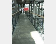 Ein moderner, überdachter Gehweg aus Beton mit Geländern aus Metall und Holz in einem Industriegebäude; links befinden sich Treppen, rechts ist eine verglaste Aufzugsanlage sichtbar. Im Hintergrund gehen zwei Personen auf den Betrachter zu.