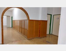 Ein schlichter Flur mit hellbraunem Holztäfelung an den Wänden, einer gewölbten Holztüröffnung und einer beige-braunen Bodenfläche. Im Hintergrund sind Türen mit grünen Rahmen sichtbar.