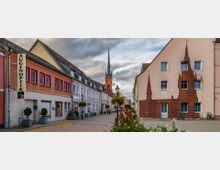 Eine Fußgängerzone mit gepflastertem Weg, gesäumt von mehrfarbigen Reihenhäusern, darunter ein Optikergeschäft. Im Hintergrund ragt der spitze Kirchturm einer Backsteinkirche in den bewölkten Himmel.