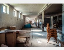 Verlassenes Gebäude mit einer langen, schmalen Halle, niedriger Decke und einer Reihe rechteckiger Fenster an der linken Wand. Die Halle ist spärlich beleuchtet und enthält gestapelte Holzstühle, Kartons, Zeltplanen und verstreute Gegenstände.