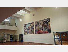 Große Innenhalle eines öffentlichen Gebäudes mit hoher Decke, sichtbaren Deckenbalken und hängenden Kugellampen. An einer hellen Wand hängen zwei großformatige, farbige Wandbilder; daneben stehen Vitrinen und Beschilderungen, rechts führt eine Glastür in einen Flur.