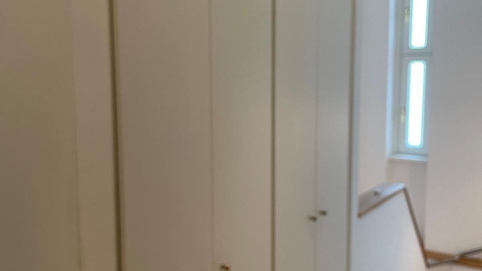 Schmaler Flur oder Treppenhausbereich mit einer langen Reihe hoher, weißer Einbauschränke entlang der linken Wand. Rechts führt eine Holztreppe mit Geländer nach unten, daneben ein schmales, hohes Fenster, das Tageslicht in den Raum lässt.