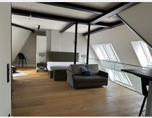 Modernes Dachgeschoss-Schlafzimmer mit sichtbaren schwarzen Stahlträgern, Holzfußboden und schrägen Deckenflächen. Ein Bett und ein Sofa stehen neben einer Glasbrüstung; mehrere Dachfenster sorgen für Tageslicht.