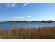 Ein See mit Schilf am Ufer im Vordergrund, im Hintergrund eine kleine Ansiedlung mit zwei Kirchtürmen und umgeben von Bäumen unter klarem, blauem Himmel.