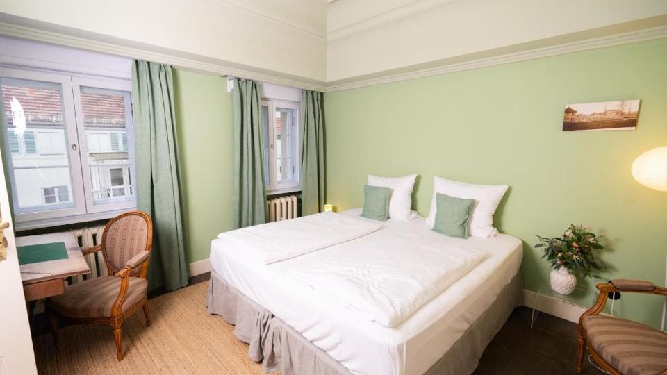 Helles Schlafzimmer mit zwei zusammengestellten Einzelbetten, grünen Wänden, hohen Stuckleisten und großen Fenstern mit grünen Vorhängen. Neben dem Bett stehen eine Stehlampe und eine Vase, am Fensterbereich ein kleiner Schreibtisch mit Stuhl sowie ein Heizkörper.