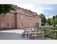 Historisches Gebäude mit orangefarbenem Putz und weißen Fenstern, umgeben von einem gepflasterten Hof mit Holzbänken und Tischen. Im Vordergrund ein Holzzaun, im Hintergrund Bäume und Wegweiser, die auf eine parkähnliche Umgebung hindeuten.