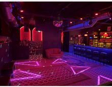 Ein Nachtclub-Interieur mit schummriger Beleuchtung in Rot- und Violetttönen. Zu sehen sind eine gepolsterte Couch, Barhocker vor einer beleuchteten Theke, geometrische Lichtprojektionen auf gemusterten Teppichen und ein Discokugel-Element an der Decke.