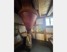 Innenraum einer historischen Mühle mit sichtbaren Holzbalken und einem rotbraunen hölzernen Trichter, der sich in einen darunter befestigten Leinensack öffnet. Im Hintergrund sind ein kleines Fenster sowie Informationstafeln an den Wänden zu sehen.