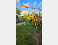 Garten mit Rasenfläche und Holz-Spielgerüst mit Schaukel und Leiter; dahinter stehen Beete mit Sträuchern und Bäumen. Im Hintergrund sind Wohnhäuser und ein blauer Himmel mit Wolken zu sehen.