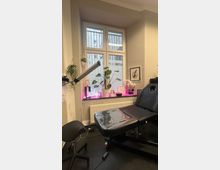 Ein heller Raum mit einer Liegefläche und einem Drehhocker, vermutlich in einem Studio oder Behandlungszimmer. Ein großes Fenster mit dahinterliegenden Gittern, einer Heizkörperbank sowie Pflanzen und Dekorationen, inklusive einer pink leuchtenden Neonlampe, prägen die Einrichtung.