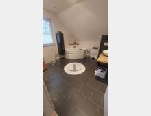 Modernes Badezimmer mit dunklem Fliesenboden und weißen Wänden. Eine Eckbadewanne mit einer abgeschrägten Decke, ein großes Fenster mit Rollladen sowie ein Teppich mit einem Ankermotiv sind zu sehen; daneben stehen dekorative Vasen und ein Wäschekorb.