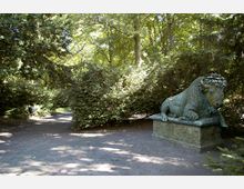 Ein Waldweg mit dichtem grünen Laubwerk, neben dem ein bronzener Wisent auf einem steinernen Sockel ruht. Der Weg führt durch einen schattigen Park mit hohen Bäumen und Büschen.