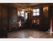 Ein historischer Saal mit hohen Decken, abgenutzten Wandverkleidungen und einem großen, goldenen Kronleuchter. An einer Wand befindet sich ein beleuchteter Barbereich, daneben sind hohe Fenster mit Gitterrahmen, die natürliches Licht hereinlassen.