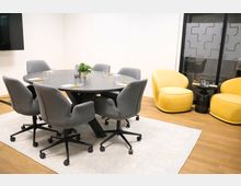 Ein moderner Besprechungsraum mit einem runden Tisch aus dunklem Holz, umgeben von sechs grauen Drehstühlen. Im Hintergrund stehen zwei gelbe Sessel neben einem kleinen Beistelltisch, und eine Glaswand mit geometrischem Muster grenzt den Raum ab.