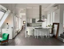 Bright open-plan loft kitchen and dining area with white cabinetry, a central island with stools, and a large metal range hood. Tall windows and slanted skylights line the left wall, with exposed hanging bulb lights, dark wood flooring, and a staircase on the right.