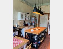 Traditionell eingerichtete Küche mit Holzmöbeln, einer zentralen Kochinsel mit Metallregalen, Hängelampen in Schwarz sowie einer Wand mit weißem Schrank und Arbeitsplatte. Im Hintergrund befindet sich ein Kühlschrank und eine Tür mit Holzrahmen.