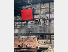 Industrielle Ruine mit rostigen Rohren, Backsteinmauerwerk und freiliegenden Metallstrukturen; eine rote Tafel mit Text ist an einer verwitterten Ziegelwand befestigt. Im oberen Bereich ist ein Wellblechdach sichtbar.