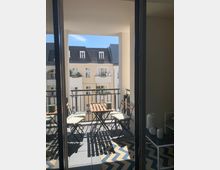 Ein kleiner Balkon mit einem Holztisch und zwei Stühlen mit weißen Sitzkissen, umgeben von einem Geländer aus Metall. Im Hintergrund sind helle Mehrfamilienhäuser mit Dachfenstern und Blumenkästen sichtbar, bei klarem, sonnigem Wetter.