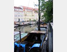 Boote liegen entlang eines schmalen Kanals vor Anker, flankiert von einem Geländer rechts. Im Hintergrund sind mehrstöckige, historische Gebäude mit roten Dächern und zahlreichen Fenstern zu sehen, die sich im Wasser spiegeln.