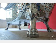 Nahaufnahme eines dekorativen Badewannenfußes in glänzender Metalloptik, gestaltet wie eine Löwentatze. Im Hintergrund sind eine bunte Mosaikverkleidung der Badewanne sowie ein roter Wandabschnitt zu sehen.