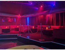 Innenraum einer Lounge oder eines Nachtclubs mit gedämpfter roter und blauer Neonbeleuchtung, niedrigen Sofas entlang der Wände und kleinen Tischen mit Kerzen. Vorne ist eine leicht erhöhte, von roten Samtseilen begrenzte Fläche mit Metallpfosten zu sehen, dahinter hängen dunkle Vorhänge.