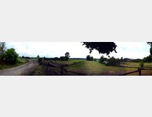 Weitläufige ländliche Szene mit grasbewachsenen Koppeln, die von Holzzäunen eingefasst sind. Im Hintergrund befinden sich Felder, Bäume und ein großes Gebäude, vermutlich ein Stall oder eine Scheune. Ein unbefestigter Weg führt entlang der Zäune durch die Landschaft.