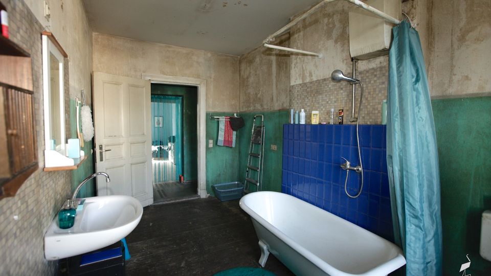 Altes Badezimmer mit freistehender Badewanne und Duscharmatur vor einer Wand aus blauen Fliesen sowie einem Waschbecken mit Spiegel links. Abgenutzte, fleckige Wände in Grün und Beige, dunkler Holzboden und eine offene Tür führen in einen angrenzenden Raum.