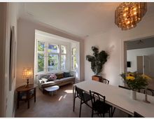 Wohnzimmer in einem Altbau mit großen, weißen Sprossenfenstern, die Tageslicht hereinlassen. Zu sehen sind eine Sitzecke mit Sofa und Beistelltisch, ein großer Esstisch mit Stühlen, eine Zimmerpflanze sowie warme Dekorationselemente wie eine Stehlampe und Blumen auf dem Tisch.