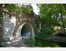 Historischer Steinbogen mit verzierter Architektur, umgeben von Bäumen und Efeu, führt über einen gepflasterten Weg und einen kleinen Wasserlauf mit einem Geländer. Ein bewaldeter Bereich und Schatten spenden eine natürliche Umgebung.