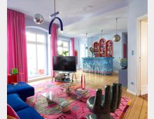 Bright, colorful living room with tall windows and pink curtains, light blue walls, and wood floors with a large patterned rug. A TV on a stand faces a blue sofa, with a glass coffee table in the center and an open bar area in the back featuring blue tiled counters and arched shelving.