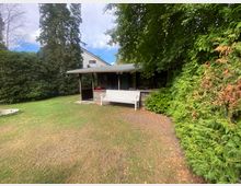 Ein Garten mit einer Rasenfläche, einem kleinen Holzpavillon mit überdachter Terrasse, einer weißen Gartenbank und umgeben von hohen Bäumen und Sträuchern. Im Hintergrund ist ein weißes Gebäude teilweise sichtbar.