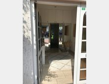 Eingangsbereich eines Hauses mit offener Glastür, hellem Fliesenboden und einem beigen Teppich. Im Inneren sind Einbauschränke, Wandspiegel und Dekorationen wie getrocknetes Pampasgras zu sehen, während im Hintergrund ein Außenbereich mit Autos erkennbar ist.