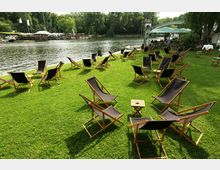 Rasenfläche am Ufer eines Flusses mit zahlreichen Liegestühlen und kleinen Beistelltischen, teils unter einem Sonnenschirm. Im Hintergrund sind Bäume sowie eine Uferpromenade mit weiteren Sitzbereichen zu sehen.