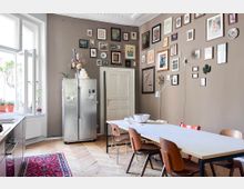 Wohnküche mit modernem Design, bestehend aus einem Esstisch mit Holzstühlen und einer Wand mit zahlreichen Bilderrahmen. Im Hintergrund befinden sich ein großer Edelstahl-Kühlschrank, eine weiße Tür sowie ein großes Fenster mit geöffneten Flügeln und Blumenkästen.