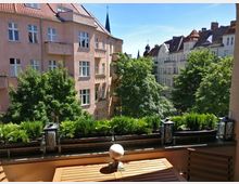 Blick von einem Balkon mit Holztisch und Stühlen auf eine von Bäumen umgebene Wohnstraße. Im Vordergrund stehen Blumenkästen mit grünen Pflanzen sowie Laternen, im Hintergrund sind mehrstöckige Altbauhäuser mit rosafarbenen und weißen Fassaden zu sehen.