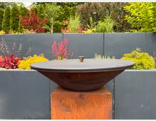 Eine runde Feuerschale aus Metall, die auf einem rechteckigen, rostfarbenen Sockel steht, vor einem bepflanzten Hintergrund mit bunten Blumen und Sträuchern sowie einer grauen, rechteckigen Wand. Im Hintergrund sind grüne Bäume und Büsche zu sehen.