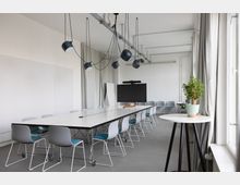 Heller, moderner Konferenzraum mit langem weißen Besprechungstisch und mehreren Stühlen auf Rollen, darüber hängende schwarze Pendelleuchten an sichtbaren Decken-Schienen. An der Stirnseite steht ein großer Bildschirm, rechts sorgen hohe Fenster mit Vorhängen und ein Pflanztopf auf einem runden Beistelltisch für Tageslicht und Akzente.