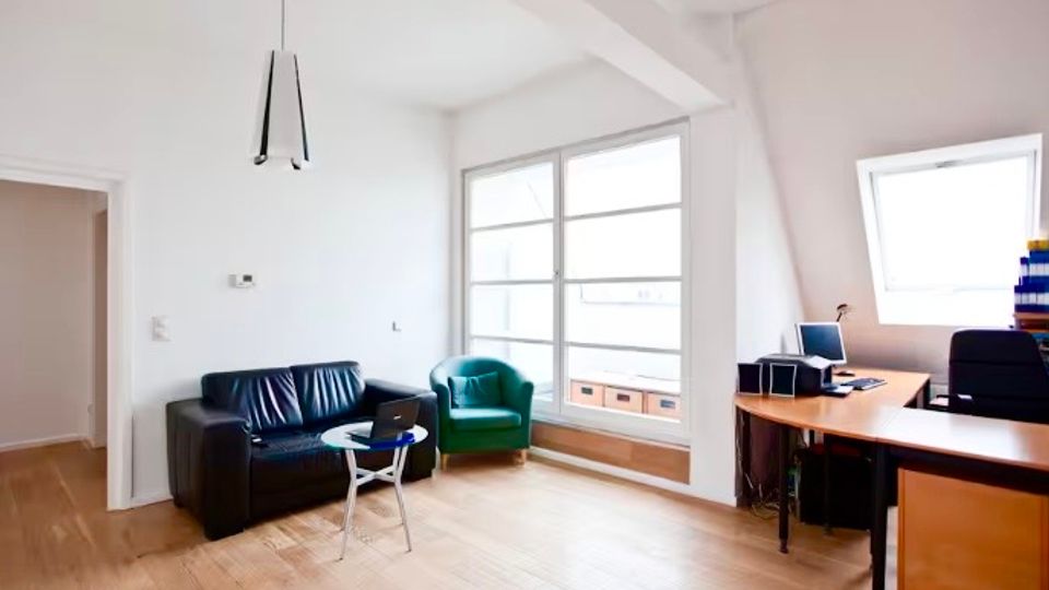 Heller Büroraum mit weißer Decke und schrägen Dachflächen, großen Fenstern und hellem Holzfußboden. Links stehen ein schwarzes Sofa und ein grüner Sessel mit kleinem Tisch, rechts ein großer Schreibtisch mit Computer.