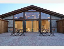 Ein modernes Gebäude mit Ziegelfassade und Holzrahmen, das durch eine große Glasfront beleuchtet ist und Einblick in einen hell eingerichteten Innenraum bietet. Vor dem Gebäude befindet sich eine gepflasterte Terrasse mit mehreren Metalltischen und Klappstühlen.