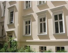 Helle Fassade eines mehrstöckigen Wohngebäudes mit mehreren weißen Fenstern, umrahmt von dezenten Verzierungen. Unten links ist ein mit "Portal 2" beschrifteter Eingang, umgeben von niedrigen Sträuchern und Pflanzen.