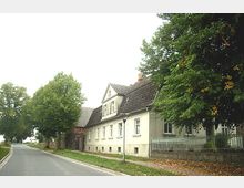 Straßenszene mit einem historischen weißen Gebäude im ländlichen Stil, flankiert von großen Bäumen. Das Gebäude hat ein Satteldach, mehrere Fenster und einen kleinen Vorgarten mit einem metallenen Zaun, während die Straße links am Haus vorbeiführt.