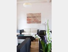 Modern eingerichteter Innenraum mit schwarzen Rattanmöbeln, beige gepolsterten Sitzflächen und Glastischen. Im Hintergrund eine weiße Wand mit einem Bild aus Ziegelstein-Optik sowie ein silberner Kerzenständer mit mehreren weißen Kerzen. Im Vordergrund sind weiße und violette Blumen zu sehen.