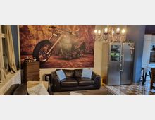 Wohnzimmer mit einer Wanddekoration, die ein großes Motorradbild zeigt. Der Raum ist möbliert mit schwarzen Ledersofas, einem Kronleuchter und einem Edelstahl-Kühlschrank, zusätzlich sind Holzboden und gemusterte Fliesen sichtbar.