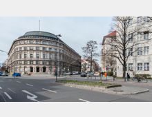 Straßenszene mit einem großen, historischen Eckgebäude mit abgerundeter Fassade und einem grünlichen Kuppeldach auf der linken Seite. Auf der rechten Seite steht ein modernes, helles Wohn- oder Bürogebäude mit vielen Fenstern; davor befinden sich Gehwege, Bäume, Fahrräder und einige Fußgänger.