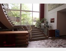 Offener Eingangsbereich eines Kinos mit einer geschwungenen Treppe, die zu einem großen Fenster mit Blick auf Bäume führt. Im Raum befinden sich Kinoplakate, eine alte Filmprojektionsmaschine und Dekorationen im Retro-Stil.