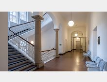 Offener Flurbereich in einem historischen Gebäude mit einer breiten Treppe und dekorativem weißen Geländer. Der Raum verfügt über helle Wände, hohe Decken, runde Hängelampen und Metallstühle entlang einer Wand.